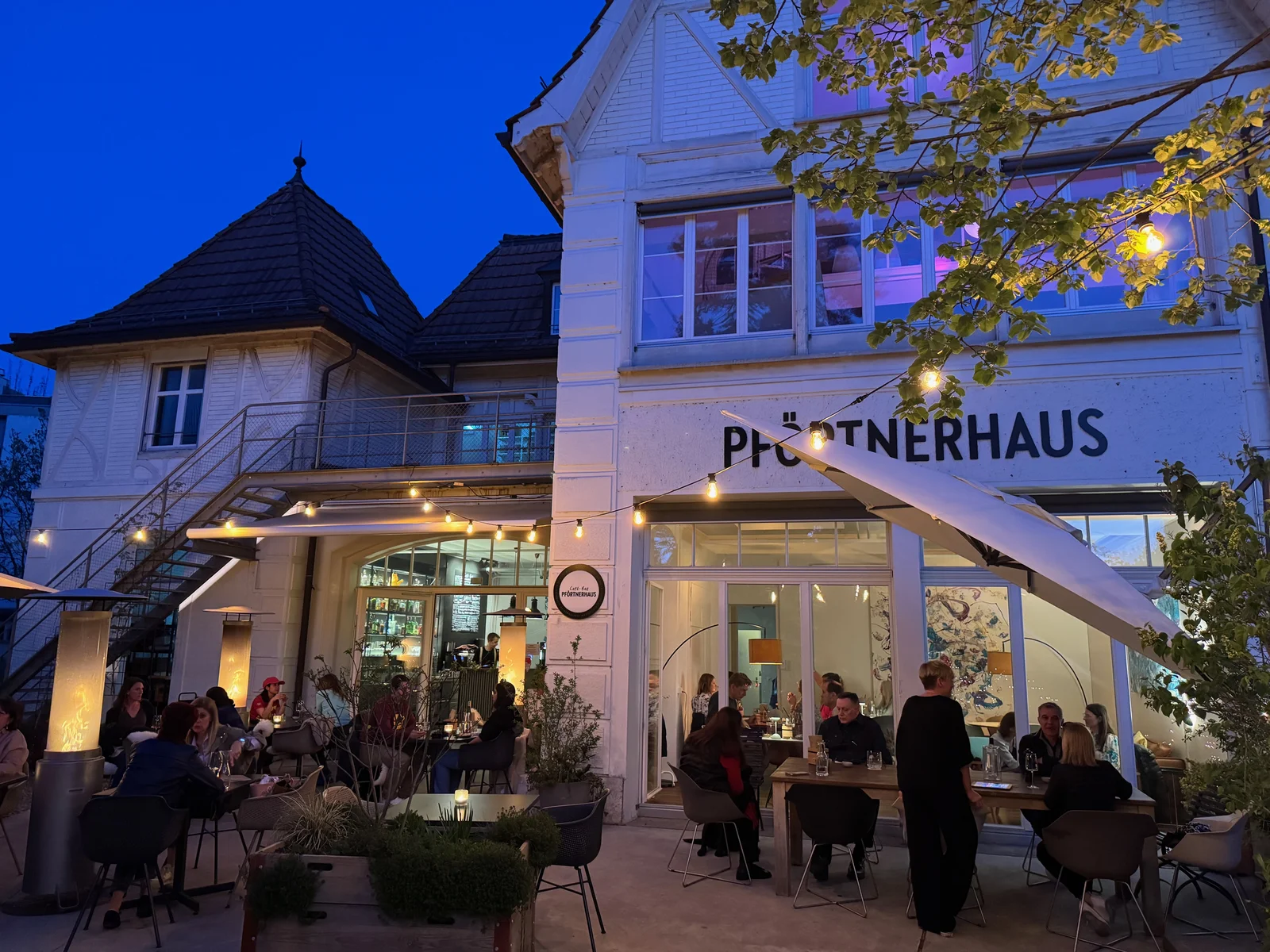 Abend-Apéro im Garten der Café-Bar Pförtnerhaus, Fassade mit Lichterkette und Gästen